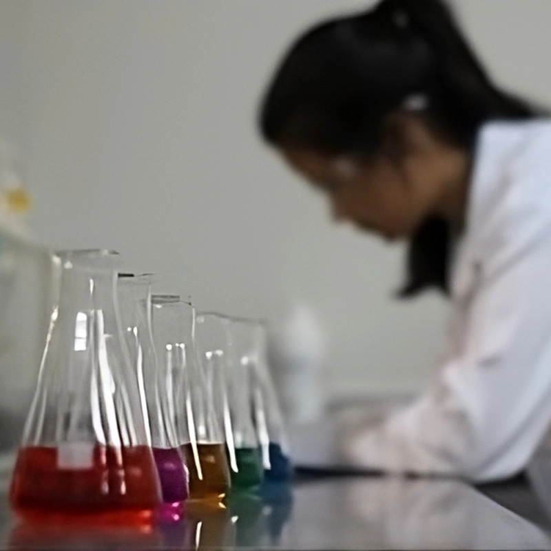 Bancada de laboratório com frascos contendo líquidos coloridos em destaque e uma pessoa desfocada ao fundo. Bancada de laboratório com frascos contendo líquidos coloridos em destaque e uma pessoa desfocada ao fundo.